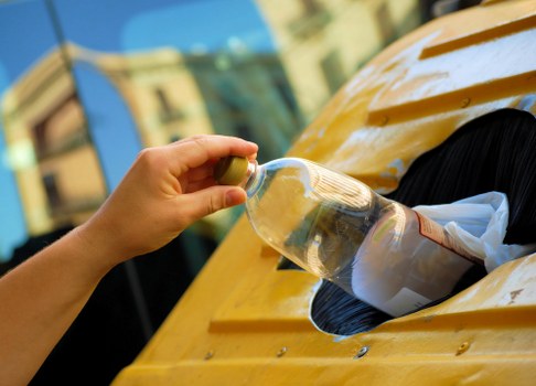 A person’s hand is seen inserting a clear plastic bottle into a yellow skip on a pavement, likely for waste disposal or collection services. The bottle appears to be a standard drinking container, with a greenish cap, and is partially filled with a liquid. The skip is made of metal, painted bright yellow, and has a rounded top with a rectangular opening through which waste is being placed. In the background, blurred elements of an urban environment are visible, including a building facade with windows and a clear sky. The setting suggests an outdoor waste removal activity, possibly associated with commercial waste services in Shortlands or nearby areas, as operated by Commercial Waste Shortlands. The scene emphasizes domestic or small-scale rubbish collection, with the focus on the action of discarding the plastic bottle into the skip, highlighting waste management practices pertinent to local rubbish removal in the Shortlands region.