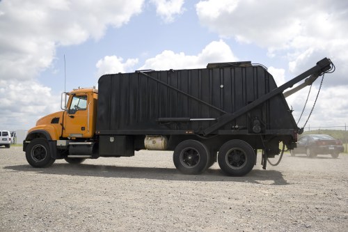 A large commercial waste collection truck with an orange cab and black enclosed waste storage body parked on a gravel surface under a partly cloudy sky. The truck features a hydraulic lift mechanism visible on the rear, used for loading and unloading refuse. The vehicle is positioned in an outdoor setting, with a few other vehicles visible in the background, including a white van and some cars. The scene suggests a waste disposal or rubbish removal context, consistent with services provided by Commercial Waste Shortlands, located in proximity to Shortlands postcode area. The image emphasizes the size and structure of the bin lorry designed for collecting commercial waste materials typical in rubbish removal operations in the local area. The overall setting highlights operational aspects of waste management vehicles used by local rubbish collection services for handling large volumes of waste efficiently.